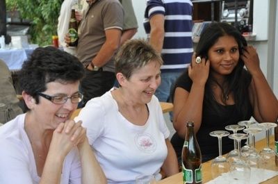 Drei Frauen sitzen an einem Tisch und lächeln und lachen. Sie sind von Weingläsern und einer Bierflasche umgeben. Im unscharfen Hintergrund sieht man eine Person mit einem Bier und eine Person mit einem gestreiften Shirt.