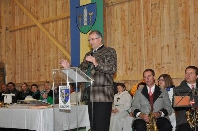 Ein Mann spricht an einem Podium in einem Raum mit Holzwänden, hinter ihm eine Flagge. Mehrere Menschen sitzen und stehen vor ihm.