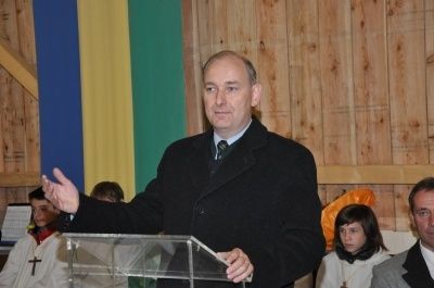 Ein Mann in einem schwarzen Anzug und Krawatte steht an einem Podium und gestikuliert mit der Hand. Dahinter ist ein farbenfroher Hintergrund und einige Personen zu sehen.