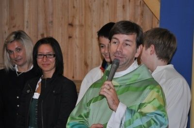 Ein Mann in einer grünen Robe spricht in ein Mikrofon, mit mehreren Personen hinter ihm, eine mit Brille und eine mit blonden Haaren.