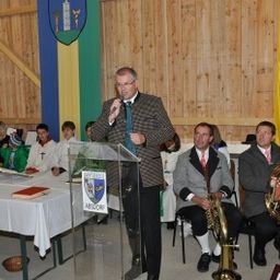 Ein Mann spricht an einem Podium mit Mikrofon vor einem Publikum, begleitet von Musikern, die Blechblasinstrumente spielen. Ein Tisch mit Büchern und einer Flagge ist im Hintergrund zu sehen.