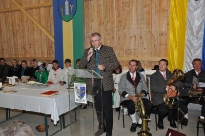 Ein Mann spricht an einem Podium mit Mikrofon vor einem Publikum, begleitet von Musikern, die Blechblasinstrumente spielen. Ein Tisch mit Büchern und einer Flagge ist im Hintergrund zu sehen.