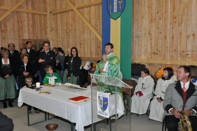 Ein Priester steht mit einem Mikrofon am Rednerpult und spricht eine Gruppe von Menschen in einem Raum mit Holzwänden an. Die Anwesenden sind Erwachsene und Kinder, einige in formeller Kleidung. Vor dem Priester steht ein Tisch mit einem Buch, einer Kerze und einer Schüssel.