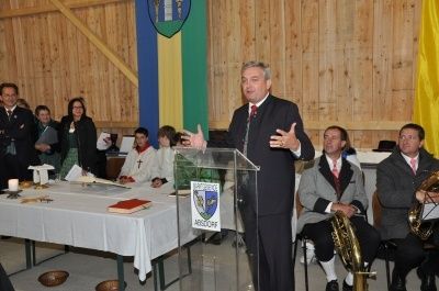 Ein Mann in einem Anzug steht an einem Podium und spricht zu einer Gruppe von Menschen in einem Raum mit Holzwänden und Flaggen.