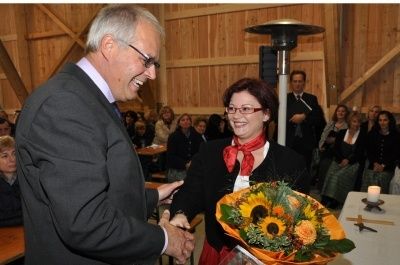 Ein Mann in Anzug und Brille schüttelt die Hand einer Frau mit rotem Schal und Brille, die einen Strauß Sonnenblumen hält, in einem Raum mit Holzwänden und mehreren zuschauenden Personen.