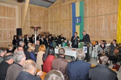 Eine Gruppe von Menschen ist in einem Scheunen für eine Veranstaltung versammelt. Ein Mann spricht an einem Podium, während andere zuhören. Eine Flagge hängt an der Wand hinter ihm.
