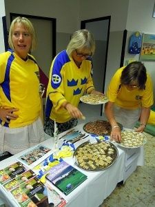Drei Frauen in gelben und blauen Uniformen bereiten Essen auf einem Tisch mit verschiedenen Tellern, Büchern und Karten zu. Eine Kerze steht in der Mitte.