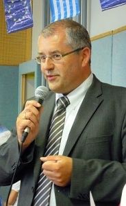 Ein Mann in Anzug und Krawatte, mit Brille, hält ein Mikrofon und spricht in dieses, während er vor einer Wand mit einer blauen Flagge steht.