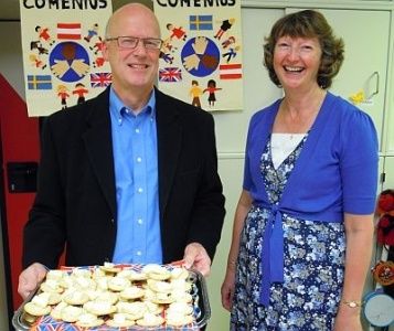 Ein Mann und eine Frau stehen nebeneinander und halten eine Tablett mit Keksen in einem Klassenzimmer. Der Mann trägt einen schwarzen Anzug und ein blaues Hemd, während die Frau ein blaues Kleid trägt. Hinter ihnen hängen bunte Poster an der Wand.