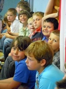 Eine Gruppe von Kindern, wahrscheinlich in einem Klassenzimmer, sitzt auf dem Boden und lächelt, einige tragen verschiedenfarbige T-Shirts, während einer ein kariertes Shirt trägt. Die Kinder scheinen etwas zu beobachten, und einer von ihnen trägt eine Halskette.
