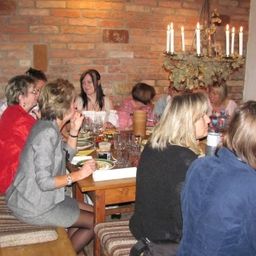 Eine Gruppe von Menschen sitzt in einem rustikalen Raum mit Backsteinwänden, einer Kronleuchter und einer Obstschale auf dem Tisch um einen Esstisch herum. Sie scheinen in Gespräche vertieft zu sein.