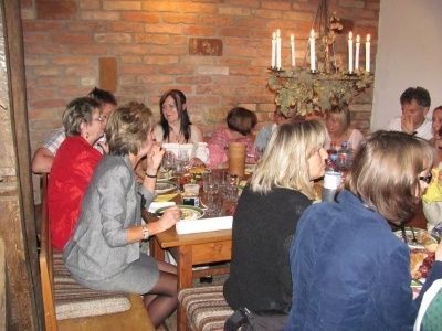 Eine Gruppe von Menschen sitzt in einem rustikalen Raum mit Backsteinwänden, einer Kronleuchter und einer Obstschale auf dem Tisch um einen Esstisch herum. Sie scheinen in Gespräche vertieft zu sein.