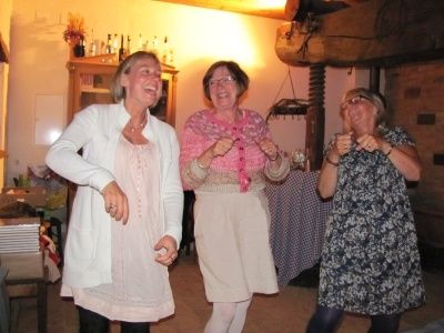 Drei Frauen tanzen in einem Raum mit Holzdecke und Regal. Eine trägt eine weiße Jacke, eine andere einen rosa Schal und die dritte ein Blumenkleid. Hinter ihnen steht ein Tisch mit einer blau-weißen Tischdecke.