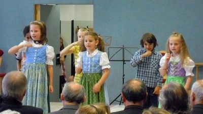 Eine Gruppe von Kindern in traditionellen Kostümen singt und tanzt vor einem Publikum. Einige halten Instrumente, andere klatschen.