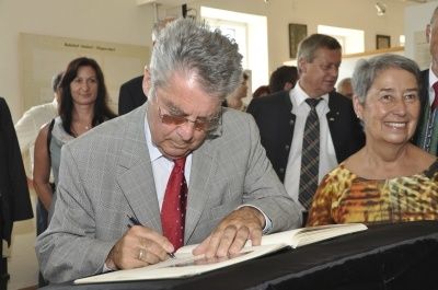 Ein älterer Mann in Anzug und Krawatte unterzeichnet ein Buch mit einem Stift bei einer Veranstaltung, während eine Frau in einem gemusterten Kleid in der Nähe lächelt.