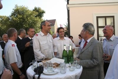Eine Gruppe von Männern in Uniformen und Anzügen steht um einen Tisch mit Essen und Getränken, möglicherweise bei einer Veranstaltung. Ein Mann spricht mit einem anderen Mann, während andere stehen und im Hintergrund sprechen.
