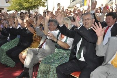 Eine große Menschenmenge in formeller Kleidung, einige in traditioneller Tracht, sitzt in Reihen und hebt die Hände, möglicherweise während einer Feier oder Veranstaltung.