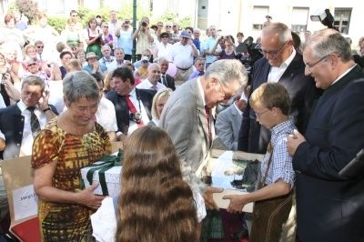 Eine große Menschenmenge versammelt sich um einen Mann mit Brille und einen jungen Jungen, die beide Boxen halten. Sie sind von anderen umgeben, darunter eine ältere Frau in einem gemusterten Kleid.