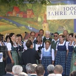 Eine Gruppe von Menschen in traditioneller Kleidung steht auf der Bühne und singt vor einem Publikum. Ein großes Banner mit der Aufschrift 'Zwei1000ele Jahre Absdorf' ist hinter ihnen angebracht.