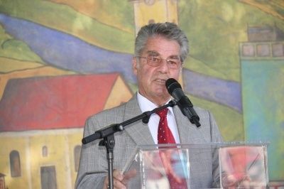 Ein älterer Mann in einem grauen Anzug und roter Krawatte steht an einem Rednerpult, hält ein Mikrofon in der Hand und spricht vor einem Publikum vor einer farbenfrohen Wandmalerei.