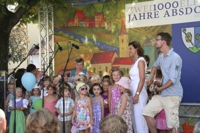 Eine Gruppe von Kindern und Erwachsenen steht auf der Bühne vor einer bemalten Kulisse. Ein Mann spielt Gitarre, und eine Frau steht neben ihm. Die Kinder tragen verschiedene Outfits, und es gibt Luftballons und ein Mikrofon auf der Bühne.
