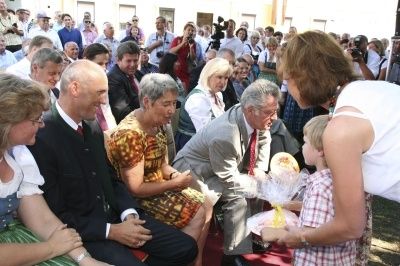 Eine Gruppe von Menschen, einschließlich Erwachsener und Kinder, ist in einem Raum versammelt. Einige von ihnen sitzen auf Stühlen, während andere stehen. Ein Mann in Anzug und Krawatte hält eine Rede, während ein junger Junge ein Geschenk hält.