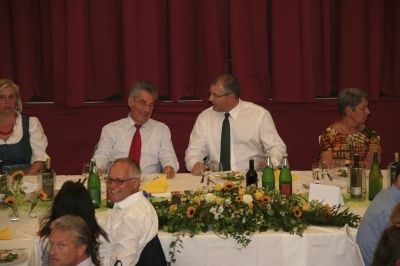 Mehrere Personen sitzen um einen langen Tisch mit Essen, Blumen und Weinflaschen. Ein Mann trägt eine rote Krawatte.