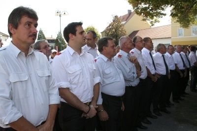 Eine Gruppe von Feuerwehrleuten steht in einer Reihe, trägt weiße Uniformen und Krawatten, vor Gebäuden.
