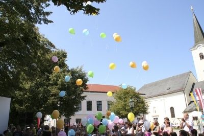 Eine Menschenmenge versammelt sich vor einer Kirche und lässt bunte Luftballons in den Himmel steigen. Bäume und Gebäude sind im Hintergrund zu sehen.