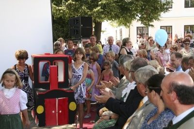 Eine Menschenmenge steht um ein Mädchen in einer Zugkostüm herum und klatscht. Ein roter und gelber Zugmodell steht vor ihr. Dahinter stehen einige Leute, und einige sitzen. Ein Gebäude steht dahinter, und ein Baum ist davor.