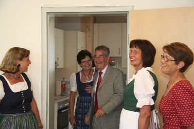 Fünf Erwachsene, gekleidet in traditioneller Kleidung, stehen in einer Küche. Sie scheinen ein Gespräch zu führen. Der Mann in der Mitte trägt eine Krawatte.