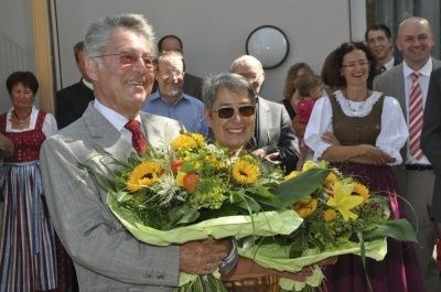 Ein Mann und eine Frau halten Blumensträuße, lächeln und stehen vor einer Gruppe von Menschen, möglicherweise bei einer Veranstaltung.