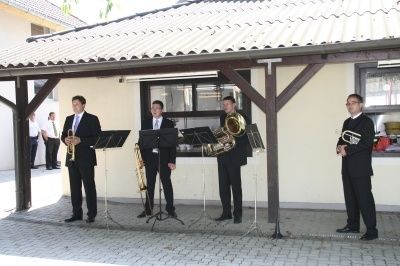 Fünf Männer in Anzügen stehen vor einem Gebäude und spielen verschiedene Blechblasinstrumente auf Ständern. Hinter ihnen ist ein Fenster mit Blick auf Menschen und eine weiße Wand.