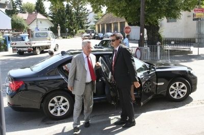 Zwei Männer in Anzügen und Krawatten stehen neben einem schwarzen Auto und besprechen möglicherweise etwas Wichtiges. Sie befinden sich auf einer Straße mit Bäumen, einem Stoppschild und einem geparkten weißen Lastwagen in der Nähe.