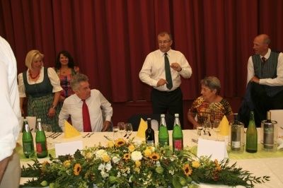 Eine Gruppe von Menschen sitzt um einen Tisch herum, mit Blumen, Flaschen und Weingläsern. Ein Mann steht und gestikuliert mit einer Krawatte. Ein roter Vorhang im Hintergrund.