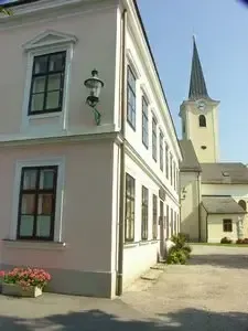 Ein weißes Gebäude mit vielen Fenstern und einem Kirchturm im Hintergrund. Vor dem Gebäude befindet sich eine Straßenlaterne und ein Blumenbeet.