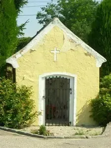Eine kleine gelbe Kapelle mit weißer Umrandung und einem Kreuz an der Vorderwand, umgeben von grünen Büschen und Bäumen.