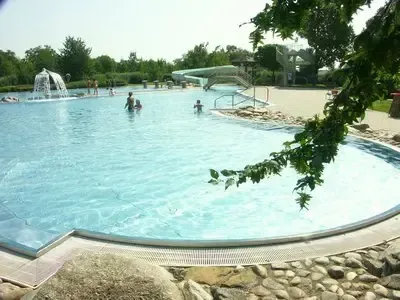 Ein Freibad mit einem Brunnen, umgeben von Felsen und Bäumen. Rechts befindet sich eine Wasserrutsche und mehrere Menschen schwimmen im Pool.