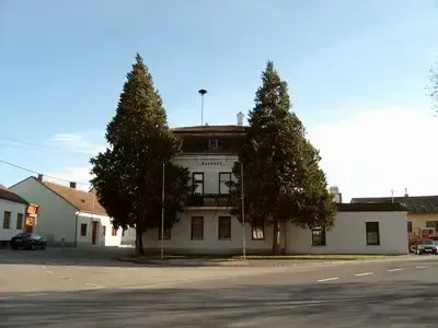 Ein weißes Gebäude mit mehreren Fenstern und zwei Bäumen davor, an einer Straßenecke gelegen. Ein Auto ist in der Nähe geparkt.