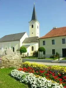 Eine weiße Kirche mit einem Turm und einer Uhr steht hinter einem farbenfrohen Blumenbeet, mit einem Gebäude mit rotem Dach in der Nähe.