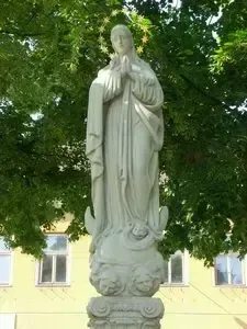 Eine weiße Statue einer Frau, wahrscheinlich die Jungfrau Maria darstellend, steht vor einem gelben Gebäude mit mehreren Fenstern. Die Statue ist von üppigem grünem Laub umgeben.