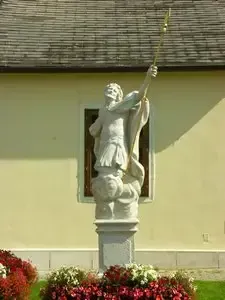 Eine Statue eines Mannes mit einem Dreizack steht vor einem gelben Gebäude mit einem Fenster. Die Statue ist von Blumen und Gras umgeben.