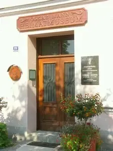 Ein weißes Gebäude mit einer braunen Holztür, über der sich ein Schild mit der Aufschrift 'Oskar Maria Graf Museum' befindet. Eine Plakette ist an der Wand neben der Tür angebracht. Ein Topf mit Blumen steht vor der Tür.