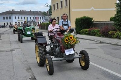 Eine Parade von Oldtimer-Traktoren mit Blumen, bei der eine Frau und ein Mann einen fahren. Dahinter folgen weitere Traktoren, Gebäude und Grünflächen.