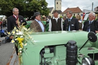 Eine Gruppe von Männern steht neben einem grünen Traktor, der mit Blumen geschmückt ist. Sie sind in Anzügen und Krawatten gekleidet und scheinen sich bei einer öffentlichen Veranstaltung zu befinden.