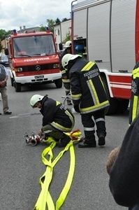 Zwei Feuerwehrleute in Uniform kümmern sich um eine verletzte Person auf der Straße, wobei ein Feuerwehrwagen in der Nähe geparkt ist.