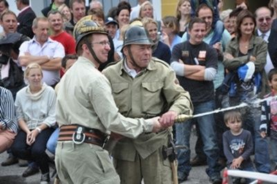 Zwei Männer in Militäruniformen schütteln sich die Hände und sprühen Wasser aus einem Schlauch vor einer Menge von Menschen.