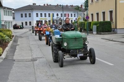 Eine Reihe von Oldtimer-Traktoren, einige mit Passagieren, fährt eine Straße hinunter, mit Gebäuden und Grünpflanzen auf beiden Seiten.