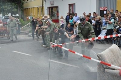 Eine Gruppe von Leuten in Uniformen zieht eine Couch mit Seilen die Straße hinunter, was eine Menschenmenge zum Zuschauen anzieht.