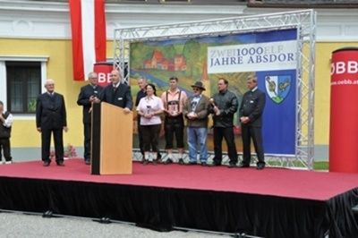 Eine Gruppe von Menschen steht auf der Bühne mit einem Banner hinter ihnen, auf dem 'Zwei1000le Jahre Absdor' steht. Auf beiden Seiten der Bühne befinden sich zwei große rote Zylinder.
